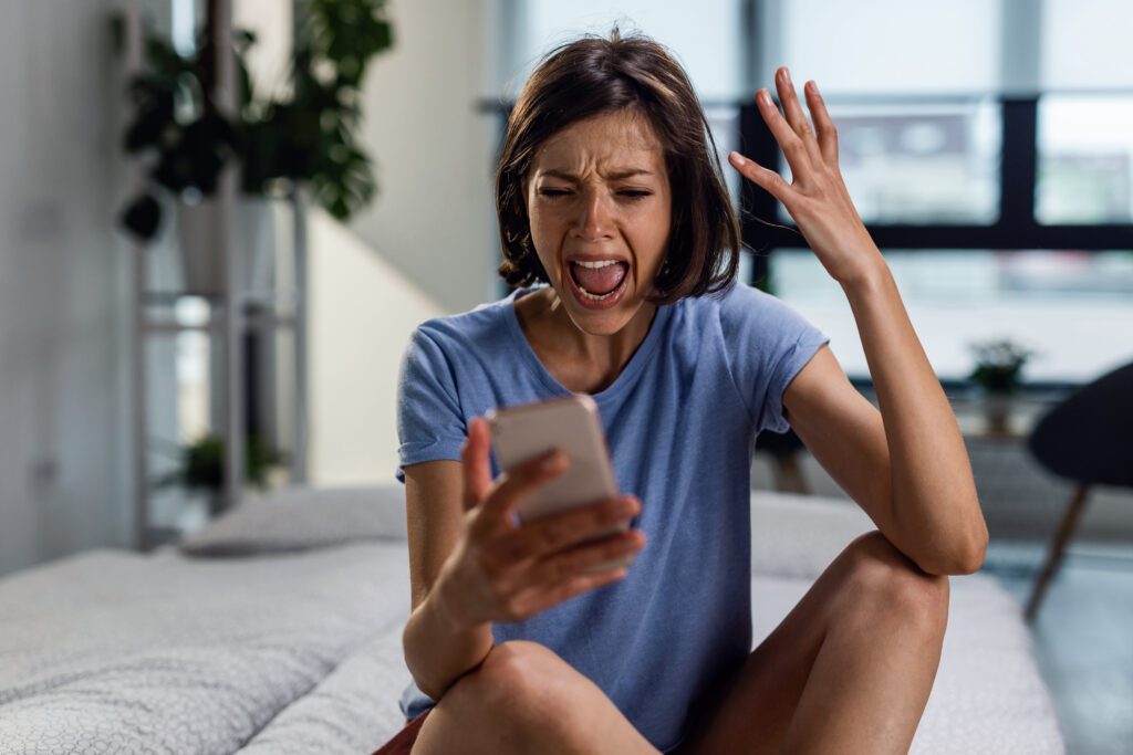 Frustrated woman shouting at her phone while sitting on the bed and reading text message she has received.