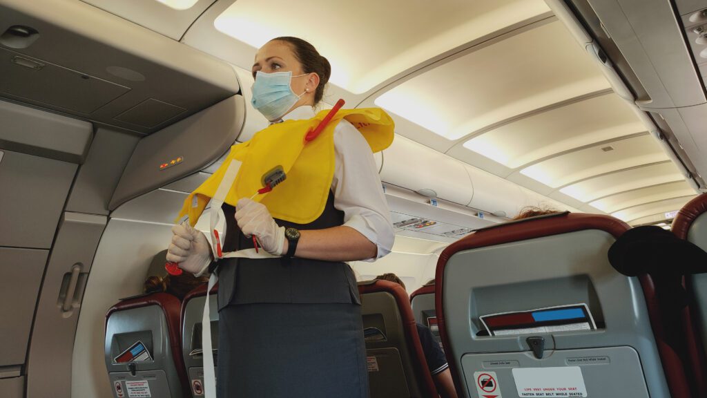 Woman stewardess airplane flight attendant in face mask presenting life vest on board before the flight departure.