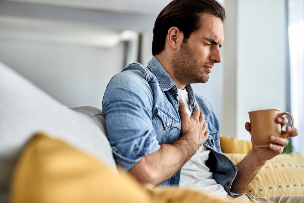 Young man feeling sick and holding his chest in pain from bad heart while drinking tea in the living room.